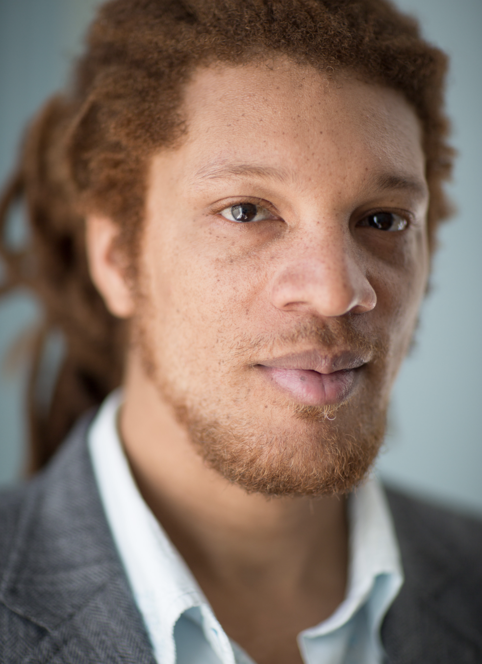 Headshot of bearded man in dreadlocks and business casual jacket and shirt, smiling slightly at the camera