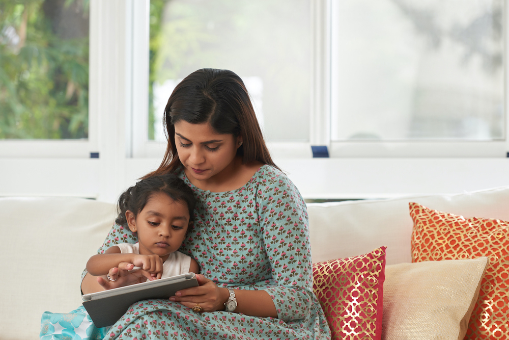 Mother and young daughter sitting on sofa, doing activities on tablet computer
