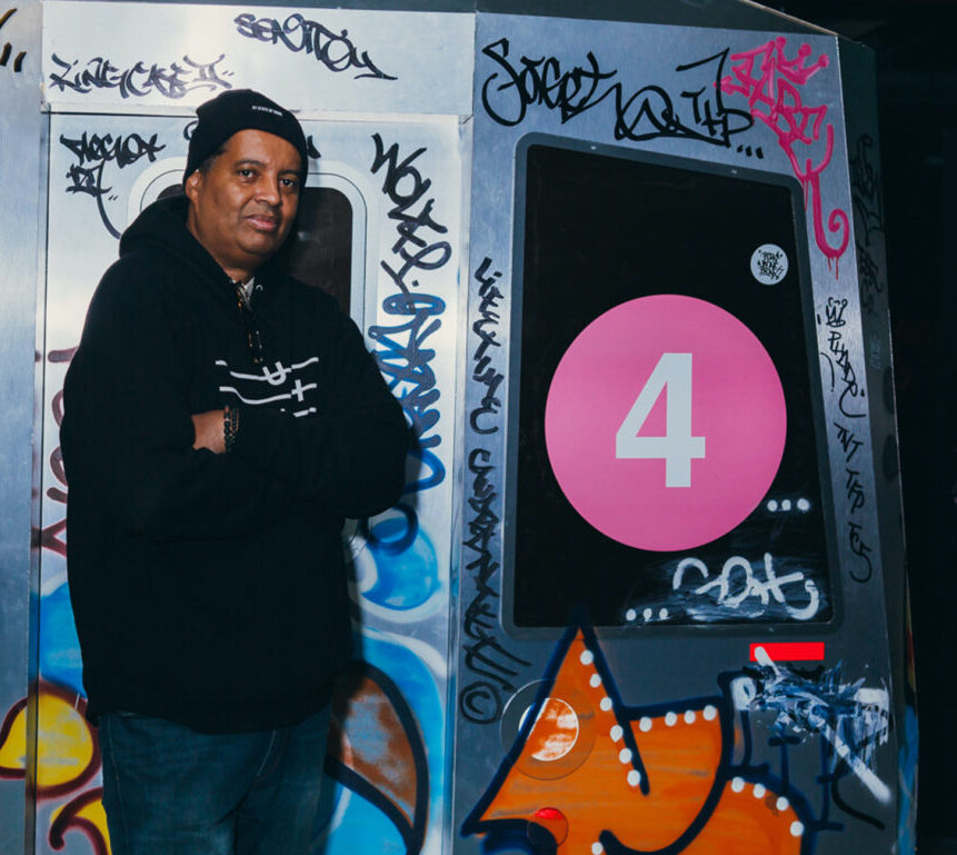 Man in sweatshirt and jeans stands, arms folded, in front of a graffiti-covered replica of a subway car