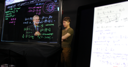 Photo of two instructors standing behind a clear lightboard that has calculations written in neon marker.