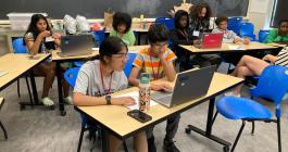 Seven students using their laptops in a classroom. One person is helping two of the students.