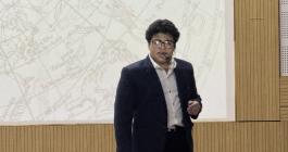 Candid photo of a teenager wearing glasses and a suit, giving a presentation in front of a map of the globe.