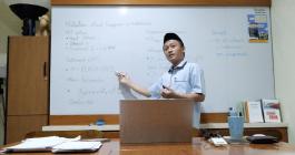 Munip Utama stands before a whiteboard, gesturing. A laptop sits open on the table in front of him.
