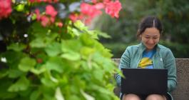 A woman on a laptop with a pink and green flowering bush in the foreground