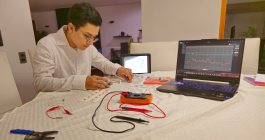A person wearing glasses and a white shirt works at a desk assembling electronic components for an electromyography tool. A laptop displaying signal data and a tablet with a schematic are visible beside measurement probes, wires, and circuit boards spread across the workspace.