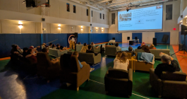 A couple dozen people sit in armchairs inside a gymnasium while watching a presentation projected on a large screen