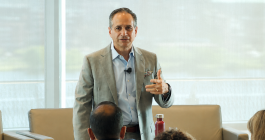 A man in tan blazer and blue shirt speaking at an event, wearing a headset microphone and gesturing to an audience, with city skyline visible through windows in the background.