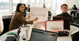 Candid photo of two individuals in an office setting during a discussion. One person is gesturing towards the whiteboard they're holding while explaining, as the other looks on attentively. Both are seated at a table with laptops and notes, surrounded by other people working. Large windows in the background suggest a bright, sunlit room.