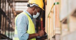 Engineer in a yellow hard hat and safety vest looks at a clipboard in a factory.