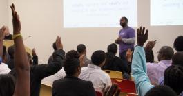 College classroom with a Black instructor standing in front of seated Black students who are raising their hands.