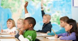 Five elementary school students at their desks, two are raising their hands