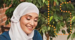 Photo of a woman in a hijab, sitting at a table and smiling at her phone.