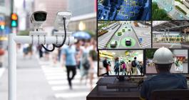 Two CCTV cameras overlook a crosswalk filled with people. A person in a hardhat looks at 6 computer screens that are showing various angles of the footage from the cameras.