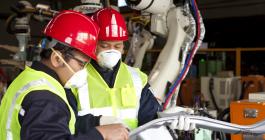 Two engineers wearing safety vests and red helmets examine a manufactured part.