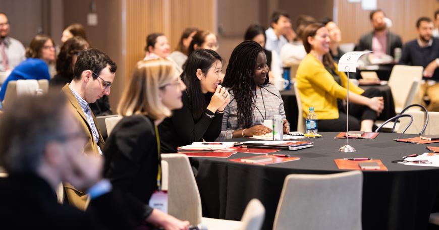Two dozen conference attendees sit around tables with papers in front of them, smiling at a speaker who is off-screen.