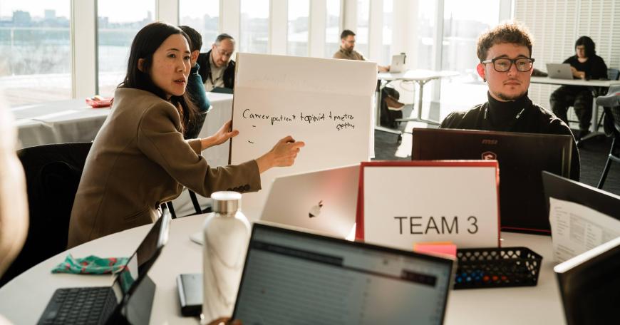 Candid photo of two individuals in an office setting during a discussion. One person is gesturing towards the whiteboard they're holding while explaining, as the other looks on attentively. Both are seated at a table with laptops and notes, surrounded by other people working. Large windows in the background suggest a bright, sunlit room.