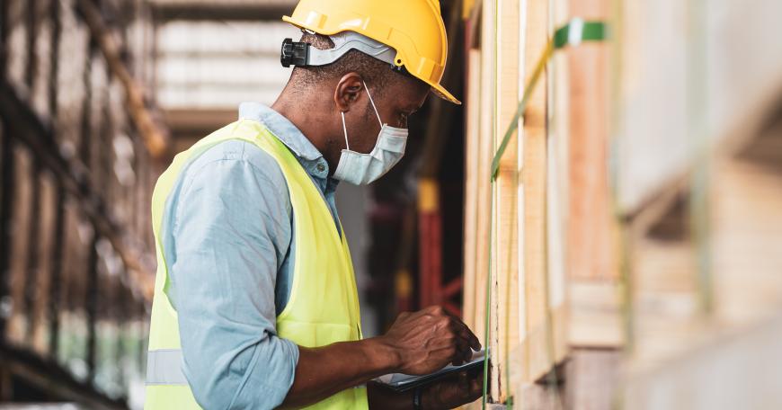 Engineer in a yellow hard hat and safety vest looks at a clipboard in a factory.