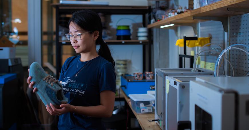 Photo of a woman in an engineering lab looking at a 3D printed sneaker with a row of 3D printers sitting on the table behind her.