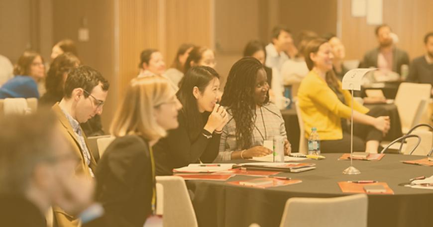 Photo of two dozen people at a conference, sitting at 5 tables and looking at a speaker who is out of frame.