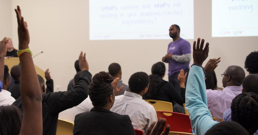College classroom with a Black instructor standing in front of seated Black students who are raising their hands.