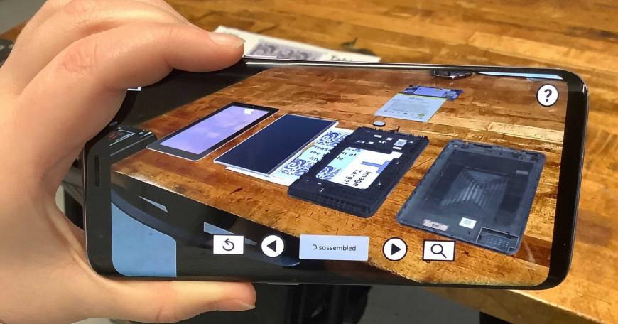 Photo of a hand holding a phone displaying augmented reality image of different tablet components as if they were on the table behind the phone. 