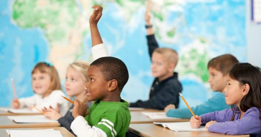 Five elementary school students at their desks, two are raising their hands