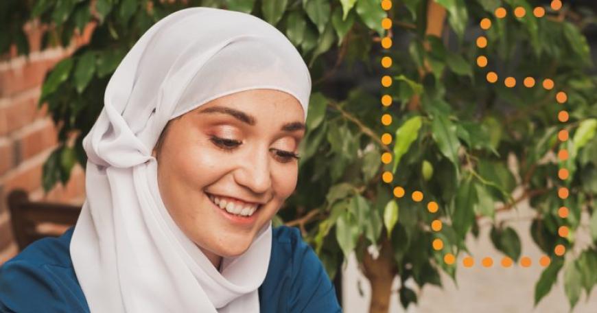 Photo of a woman in a hijab, sitting at a table and smiling at her phone.