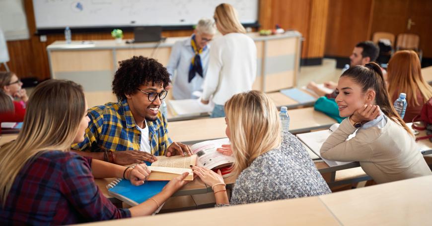 Group of 4 college students engaged in conversation while professor helps one student