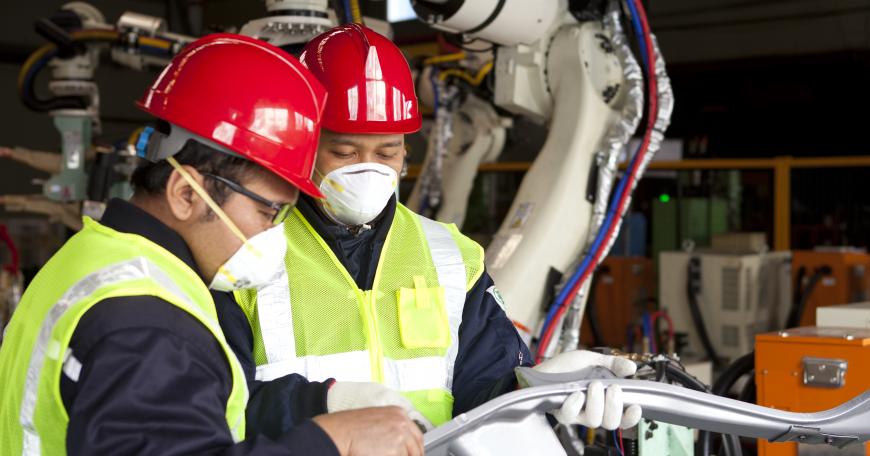 Two engineers wearing safety vests and red helmets examine a manufactured part.