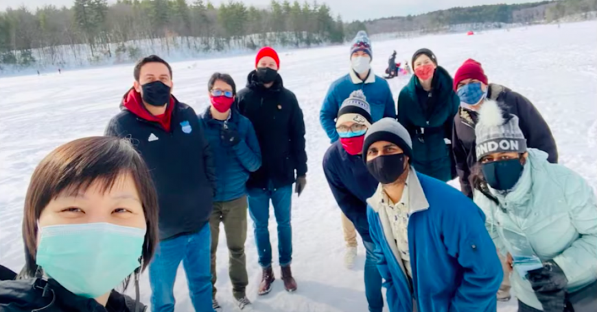 Multiethnic group of men and women dressed in winter wear and face masks, standing on a frozen lake.