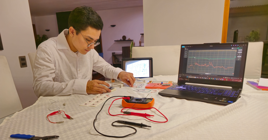 A person wearing glasses and a white shirt works at a desk assembling electronic components for an electromyography tool. A laptop displaying signal data and a tablet with a schematic are visible beside measurement probes, wires, and circuit boards spread across the workspace.