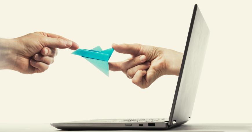 Photo of hand sticking out of a laptop to pass origami rocket to someone else.