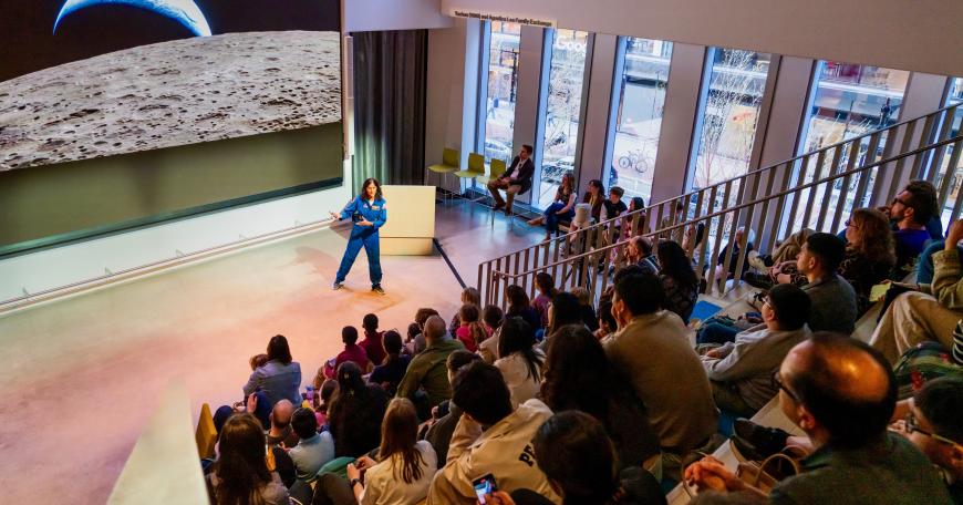 A large crowd watches Suni Williams in a NASA flight suit with a photo of Earth of space on the screen behind her.