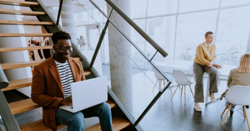 Photo of worker sitting on a staircase while typing on laptop. In the background, two coworkers are in conversation at a table.