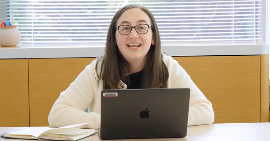 Photo of a seated woman with glasses smiling behind an open laptop.