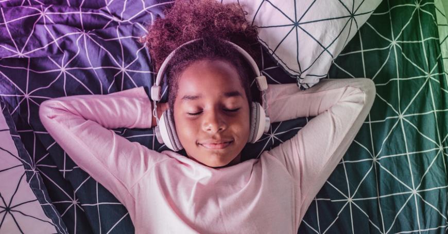 Photo of an elementary school-aged girl laying down with her eyes closed and her arms resting behind her head, wearing headphones.