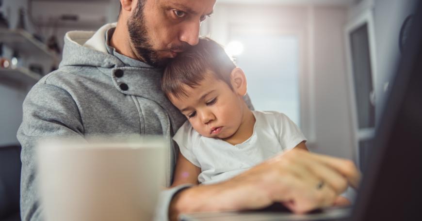 Father kisses the head of sleeping child while working on a laptop