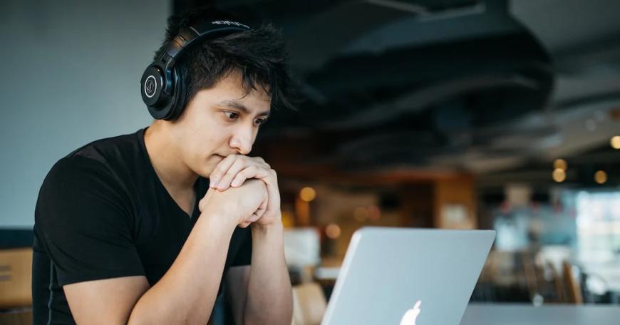 Photo of a man wearing over-ear headphones sitting at a table with his hands clasped together. He's staring at an open laptop in front of him.