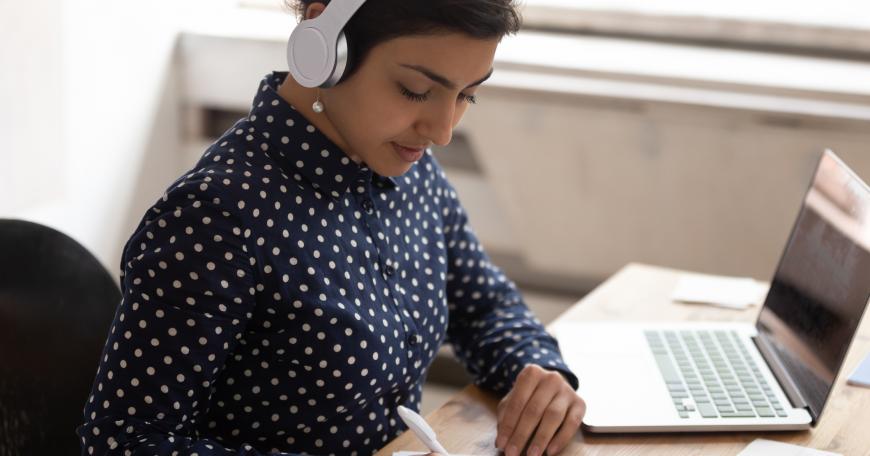 Young woman sitting at laptop, wearing headphones, taking notes