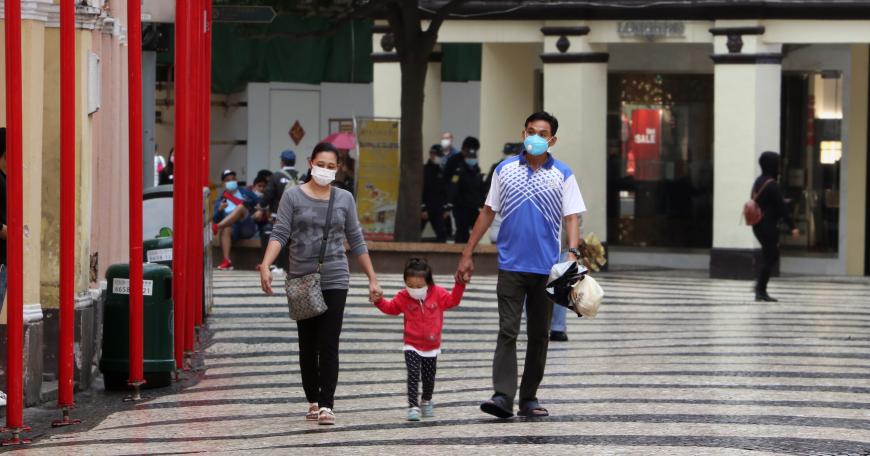 A couple hold the hands of their young child as the three, all masked, walk through an outdoor shopping center.