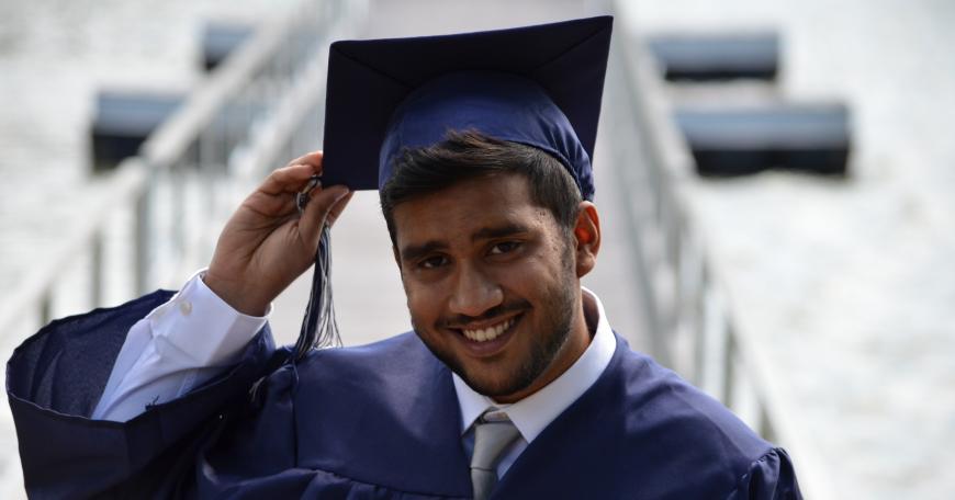 Young bearded man in graduation cap and gown smiles at the camera.