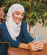 Photo of a woman in a hijab, sitting at a table and smiling at her phone.