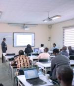 Candid photo of a classroom with over a dozen adults seated at desks with open laptops, while an instructor stands at the front to explain the lesson displayed on the screen on the wall.