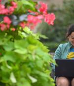 A woman on a laptop with a pink and green flowering bush in the foreground