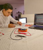 A person wearing glasses and a white shirt works at a desk assembling electronic components for an electromyography tool. A laptop displaying signal data and a tablet with a schematic are visible beside measurement probes, wires, and circuit boards spread across the workspace.