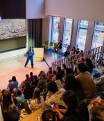 A large crowd watches Suni Williams in a NASA flight suit with a photo of Earth of space on the screen behind her.