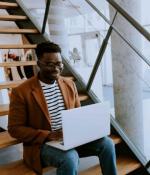 Photo of worker sitting on a staircase while typing on laptop. In the background, two coworkers are in conversation at a table.