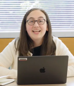 Photo of a seated woman with glasses smiling behind an open laptop.