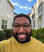 Photo of a smiling man wearing glasses, standing outside by flower bushes and houses.