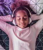 Photo of an elementary school-aged girl laying down with her eyes closed and her arms resting behind her head, wearing headphones.